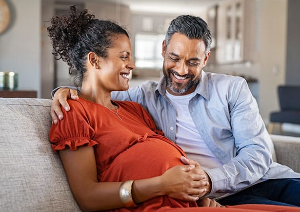 Pregnant woman and man sit on couch smiling at eachother